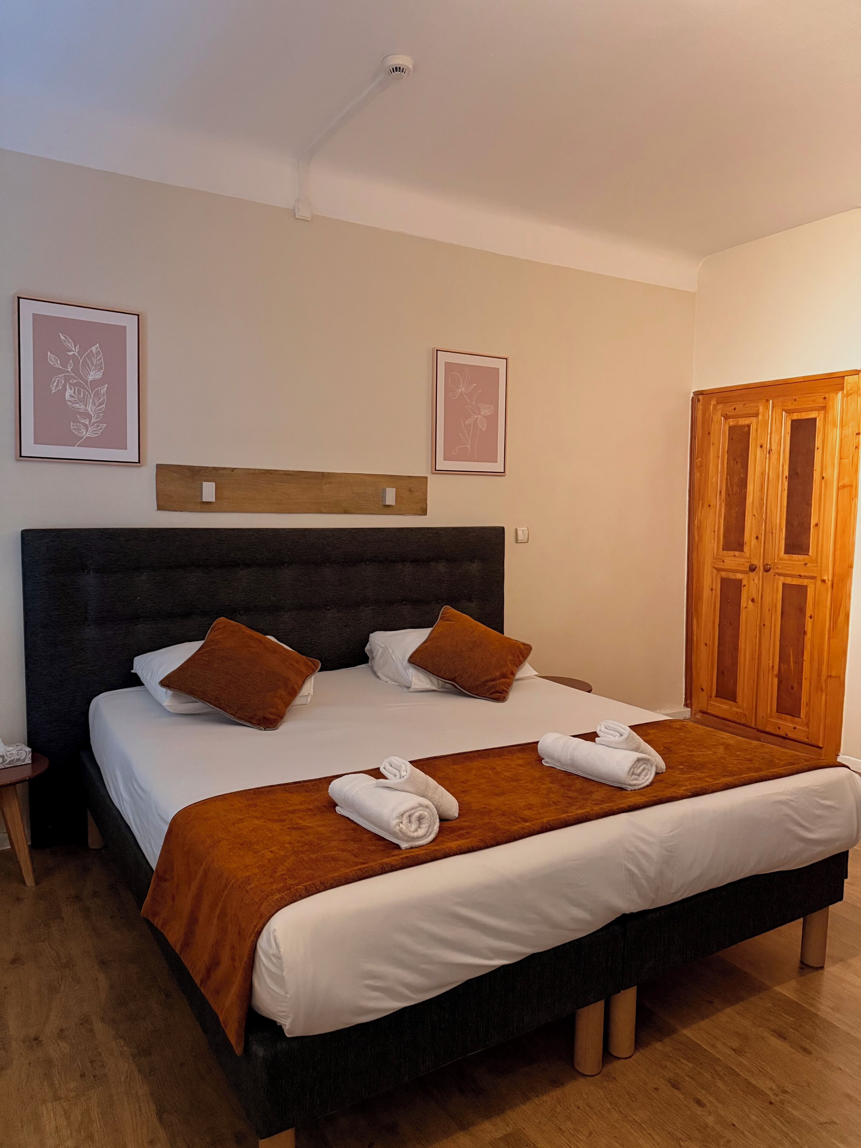 A neatly made bed with a dark headboard, two pillows, a brown throw blanket, and two rolled towels on a wooden bedside table.