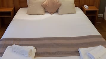 Un lit bien fait, avec des oreillers blancs et beiges, une tête de lit en bois et deux tables de chevet avec des lampes. Un tableau encadré est accroché au-dessus du lit.