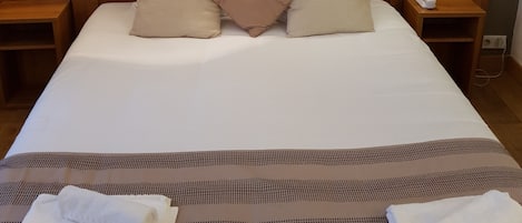 Un lit bien fait, avec des oreillers blancs et beiges, une tête de lit en bois et deux tables de chevet avec des lampes. Un tableau encadré est accroché au-dessus du lit.