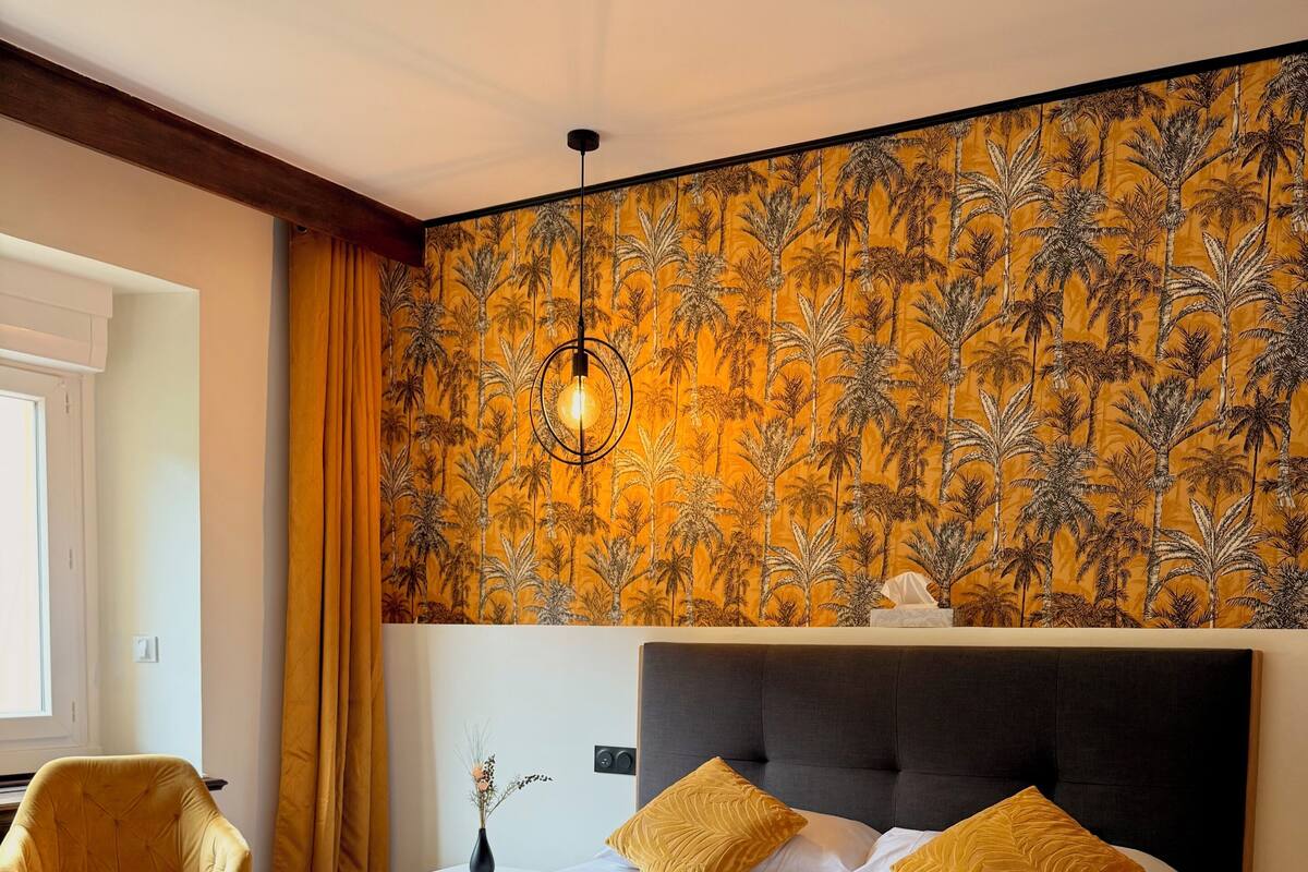 Une chambre d’hôtel avec un grand lit, une tête de lit, une table de chevet, une chaise et un mur orné d’un motif tropical.