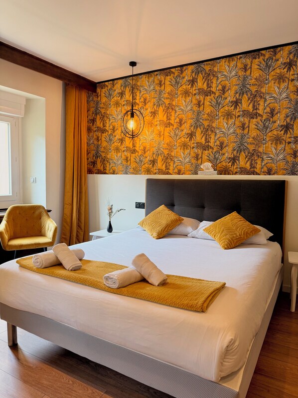Habitación de hotel con una cama grande, cabecera, mesita de noche, una silla y una pared con estampado tropical.