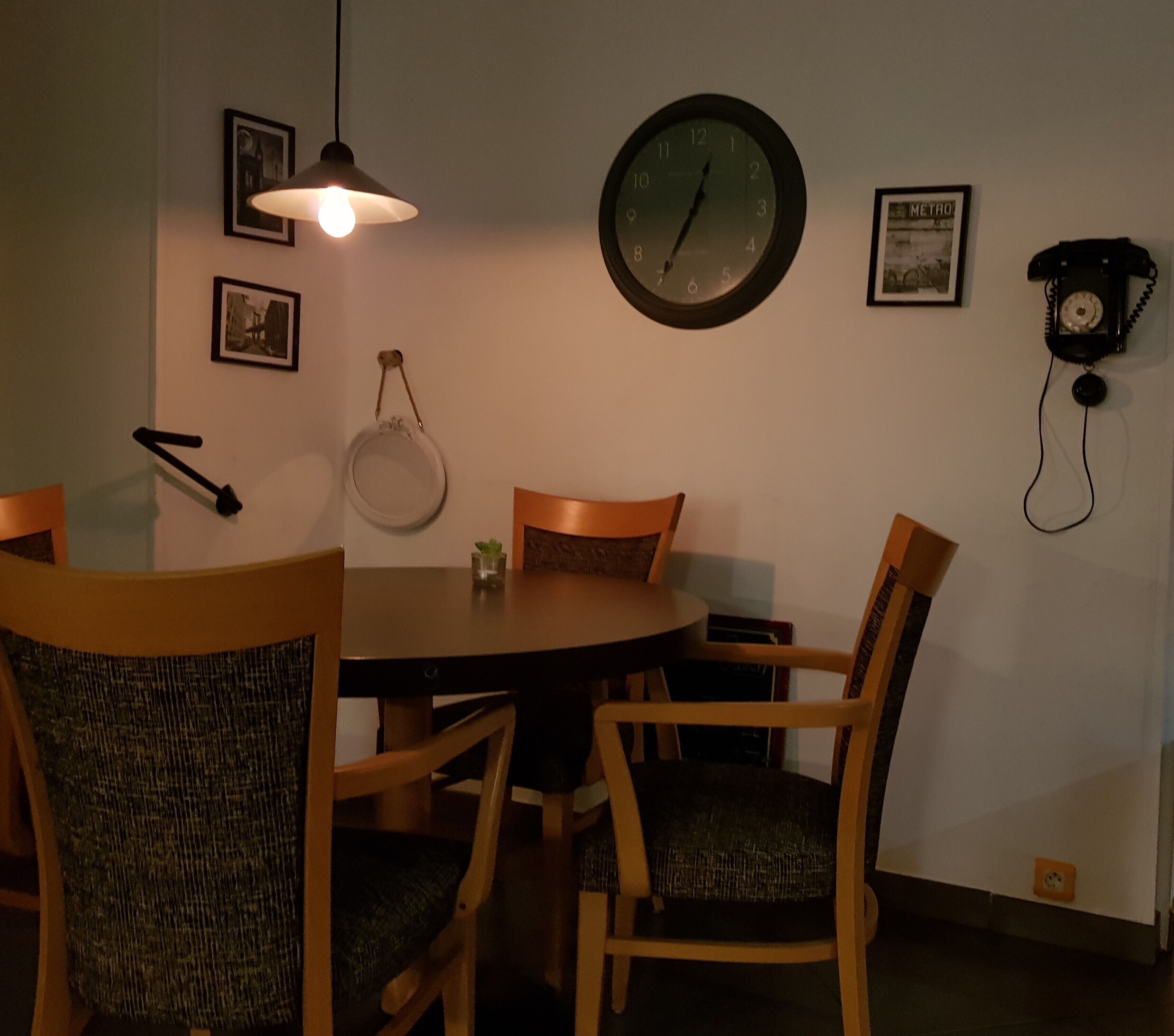A dining area with a round table, four chairs, a wall clock, a hanging lamp, and a vintage rotary phone.