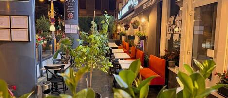 Un área de comedor al aire libre con mesas y sillas, plantas en macetas y un letrero para un restaurante.