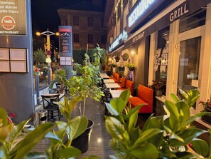 Un área de comedor al aire libre con mesas y sillas, plantas en macetas y un letrero para un restaurante.