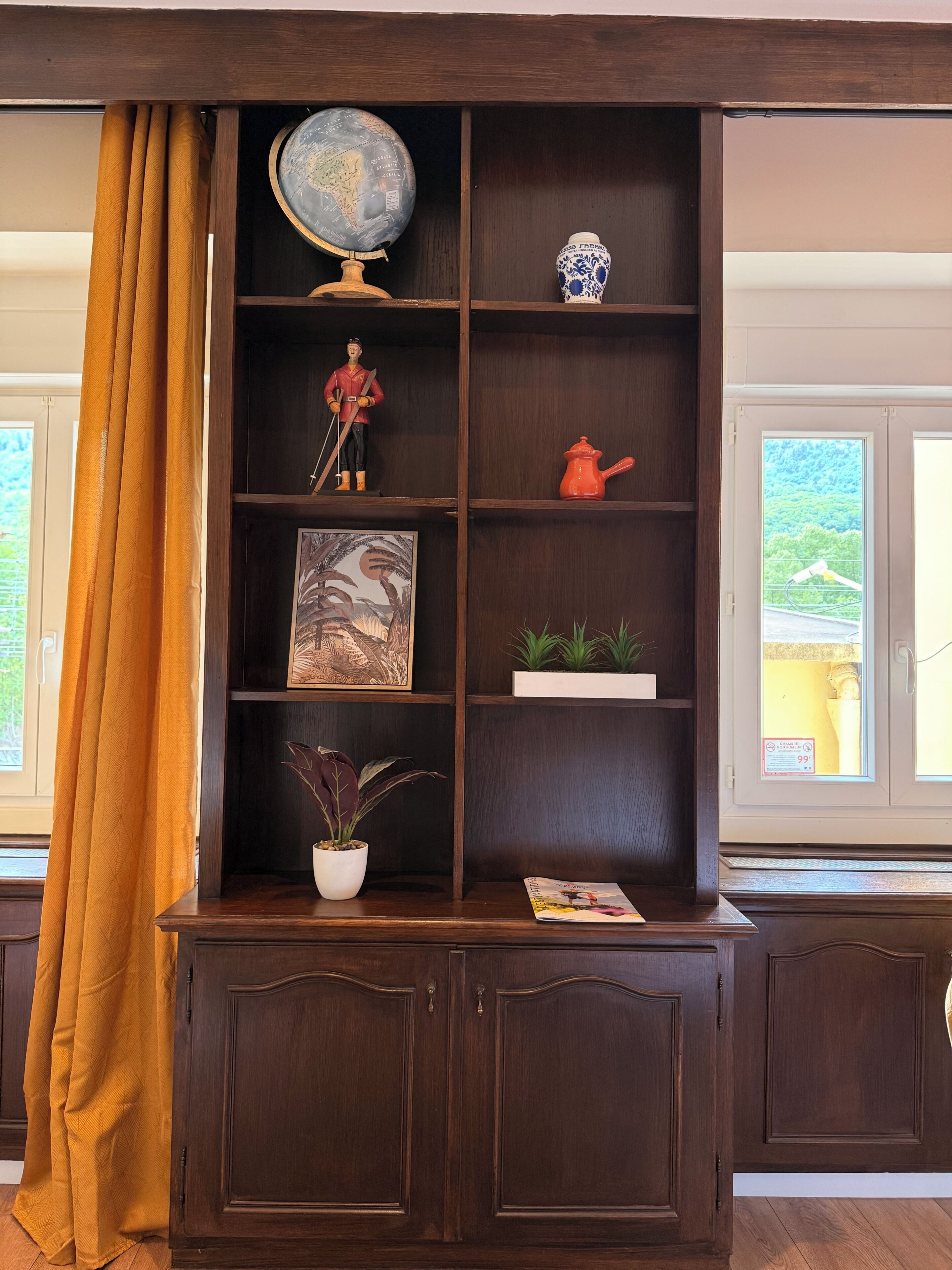 A wooden bookcase with a globe, a figurine, a framed picture, a potted plant, and a red teapot.