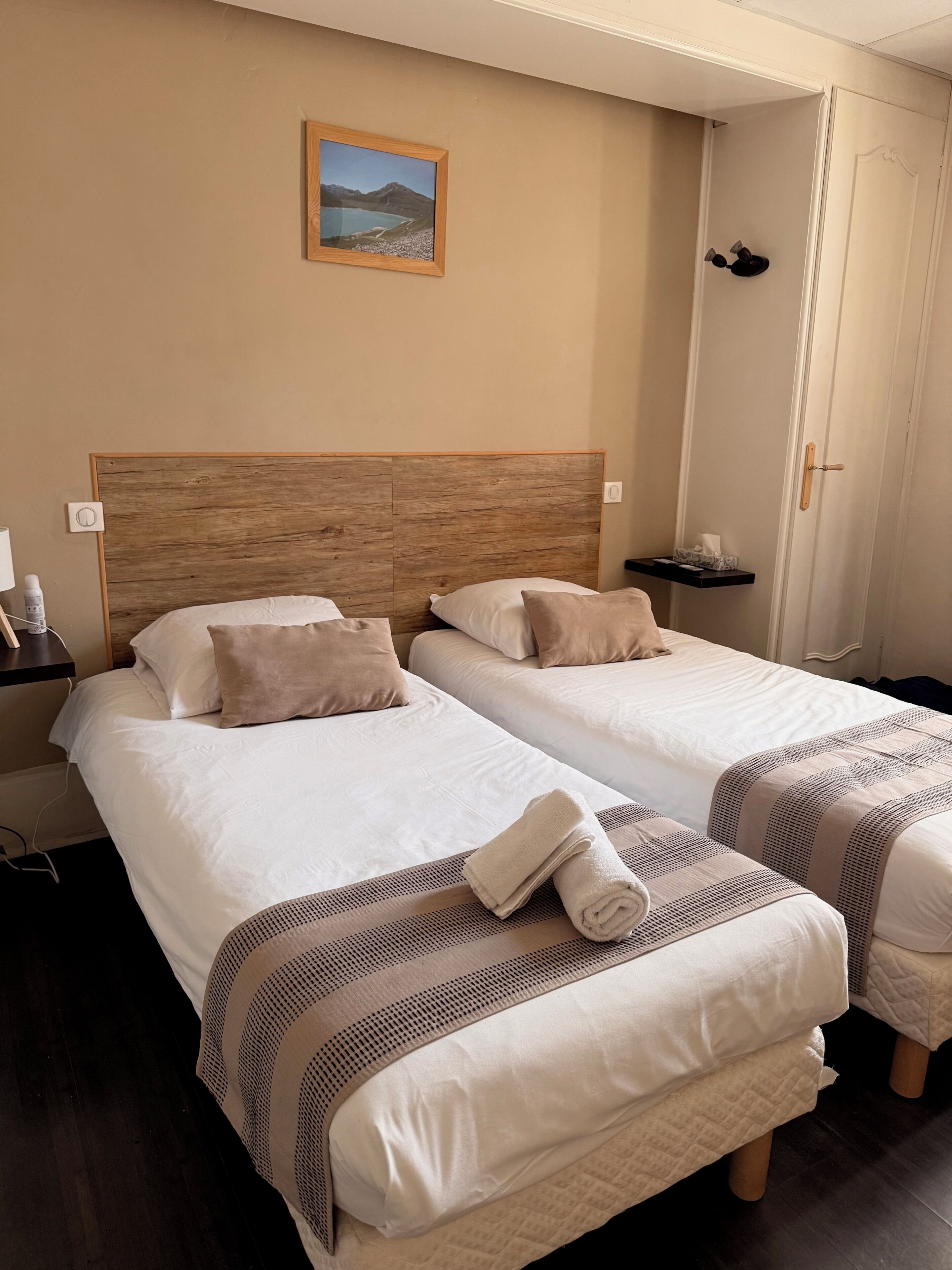 A hotel room with two beds, a wooden headboard, a painting on the wall, a nightstand, and a door.