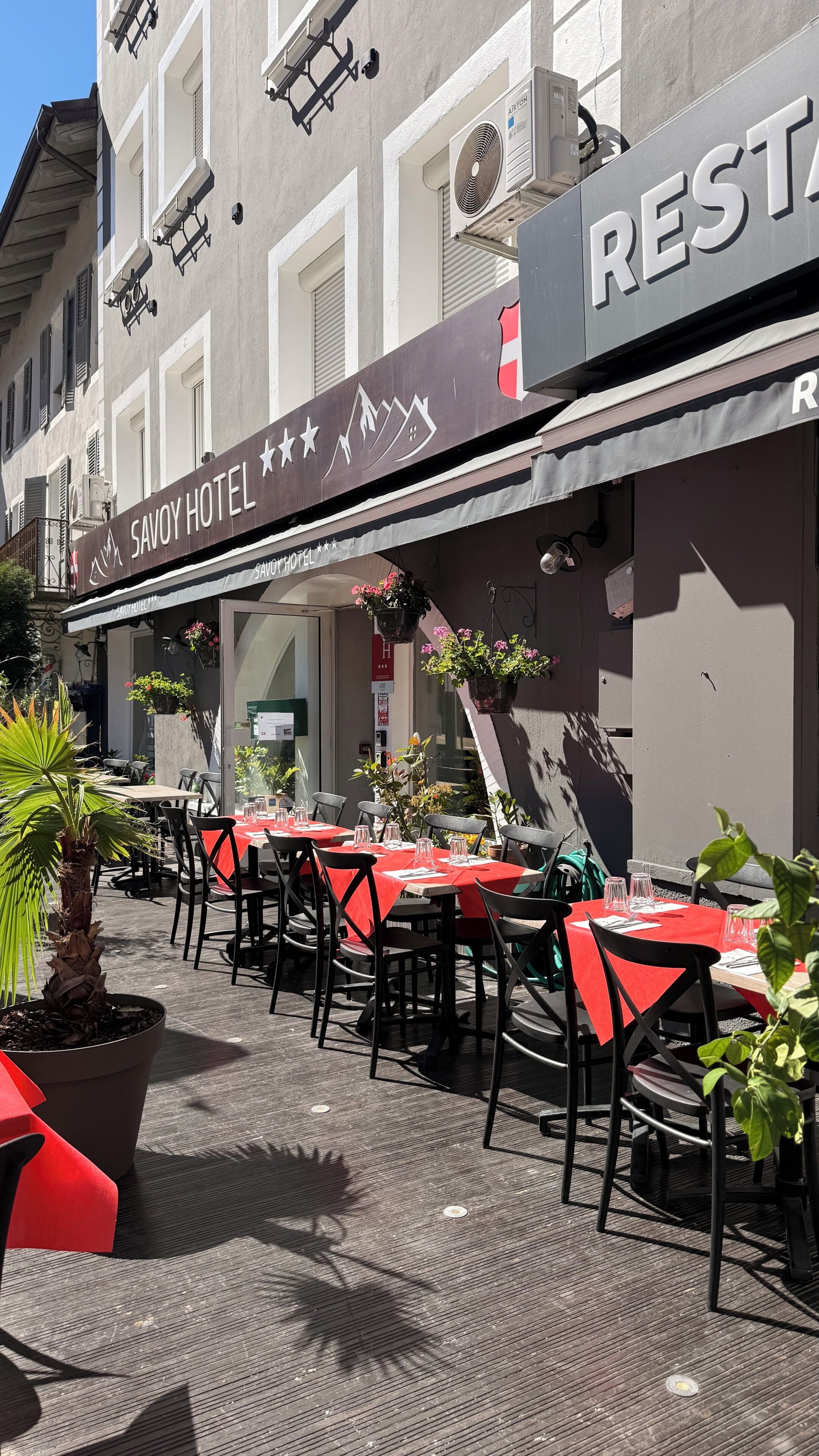 An outdoor seating area of a hotel with tables and chairs, a sign for the hotel, and hanging flower baskets.