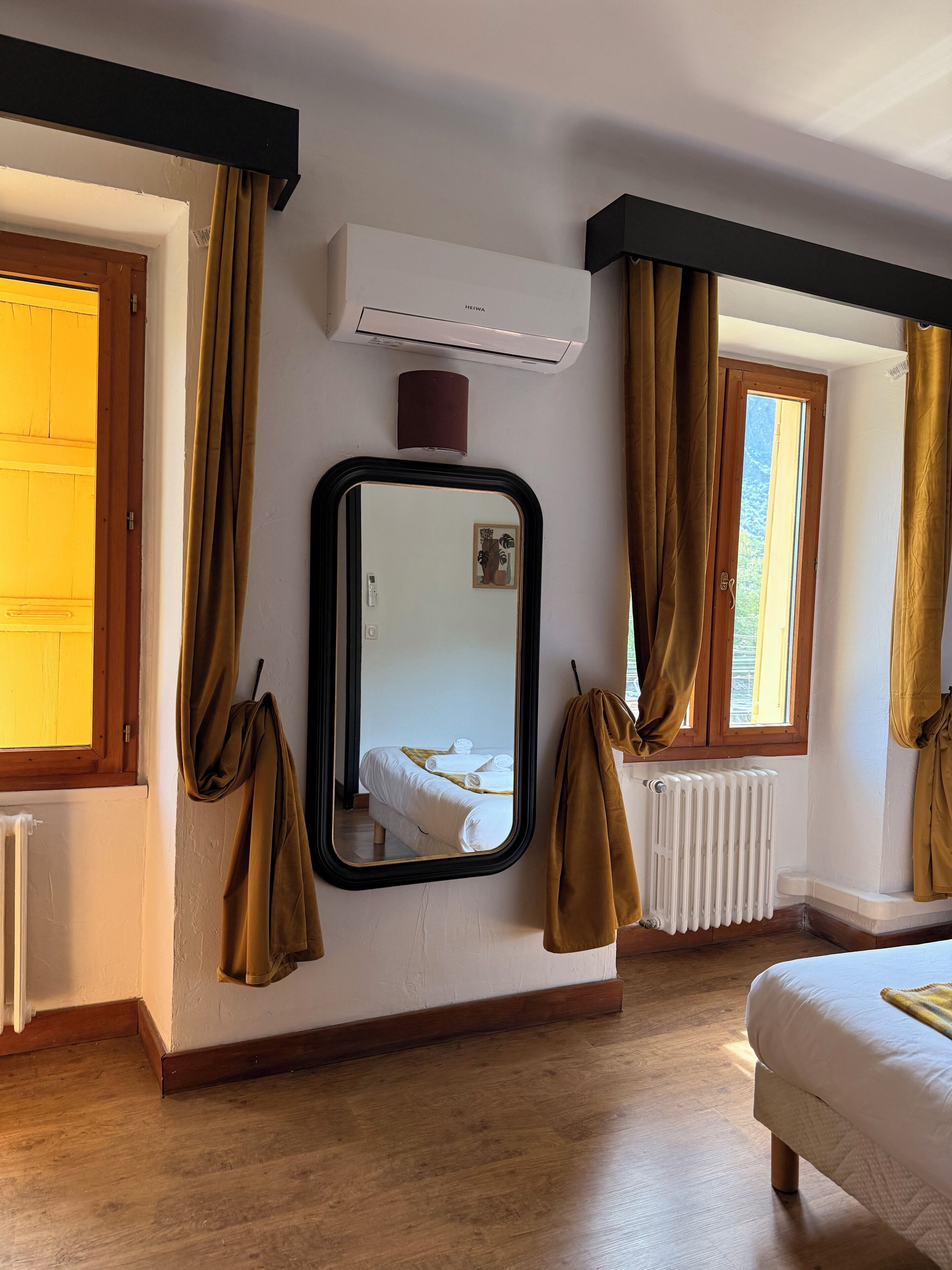 A hotel room with a bed, a large mirror, an air conditioning unit, a radiator, and a window with curtains.