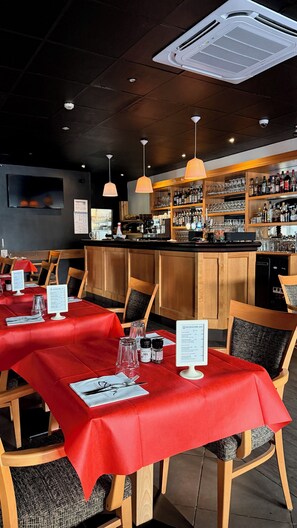Interior de restaurante con manteles rojos, sillas de madera, iluminación colgante, una zona de bar con estantes llenos de botellas y un atril para el menú.
