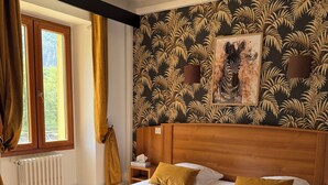 Habitación de hotel con una cama grande, cabecera de madera, ventana con cortinas, radiador y pared con estampado de hojas tropicales.