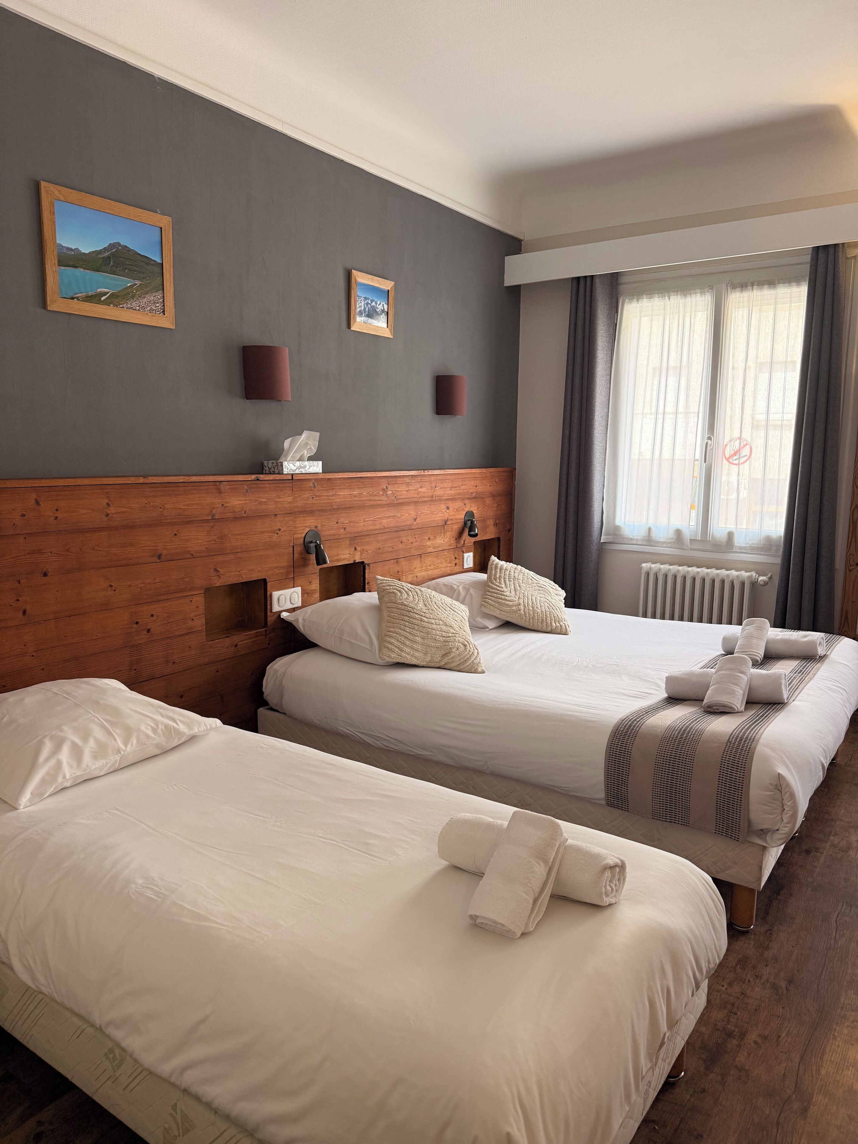 A hotel room with two beds, a wooden headboard, a window with curtains, and two wall-mounted lamps.