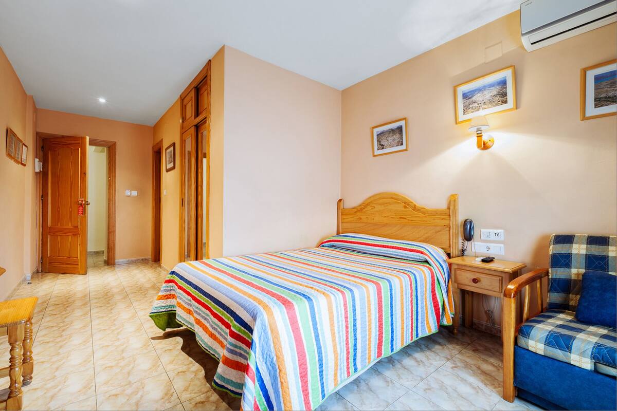 Ein Schlafzimmer mit einem Bett, einem Nachttisch aus Holz, einem Sessel und einem an der Wand befestigten Fernseher.