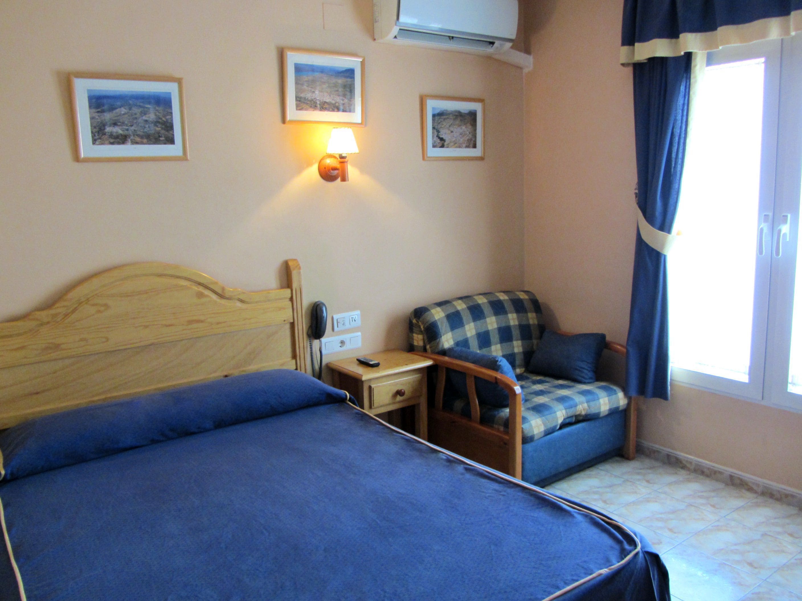 Habitació d'hotel amb dos llits, una taula petita amb una làmpada, una cadira i una finestra amb cortines.