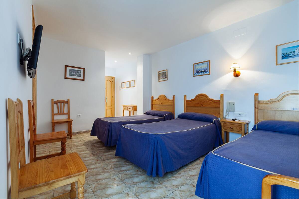 Ein Hotelzimmer mit drei Betten, einem Holzstuhl, einem kleinen Tisch und einem an der Wand befestigten Fernseher.