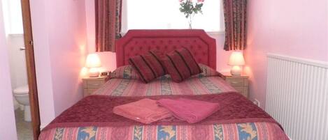 A bedroom with a large bed, two bedside tables with lamps, a window with curtains, and a radiator.