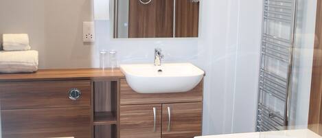 Un baño moderno con un lavamanos blanco, tocador de madera, inodoro y ducha con mampara de vidrio.