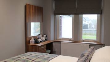 Un dormitorio con cama, escritorio con espejo, radiador y ventana con vistas a casas.