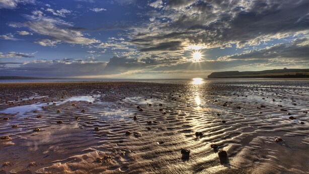 Une plage de sable humide, parsemée de rochers, sous un ciel partiellement nuageux, avec le soleil se couchant ou se levant.