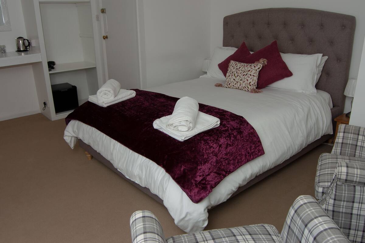 A neatly made bed with a white comforter and a dark red blanket, a grey upholstered headboard, and a bedside table with a lamp.