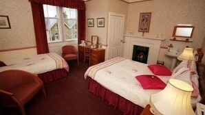 A hotel room with two beds, a desk, a chair, a fireplace, and a window with curtains. - Bellevue Guest House (Kelso)