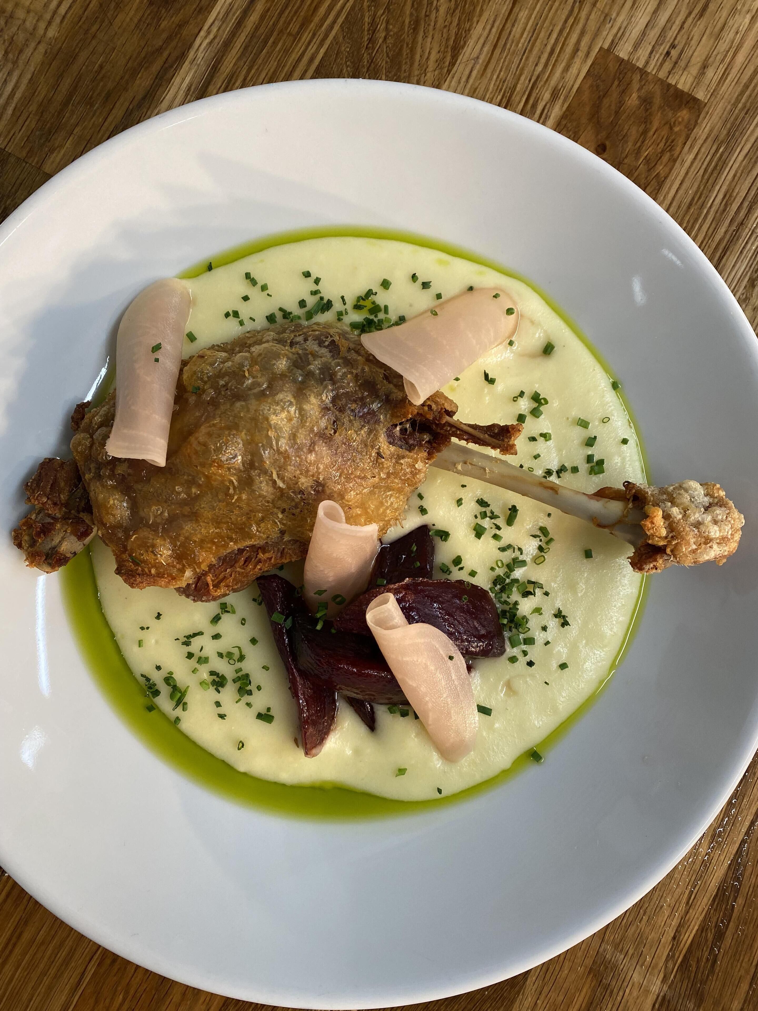 Ein weißer Teller mit einem gebratenen Entenkeule, einer Brotscheibe und grüner Sauce, garniert mit Schnittlauch.