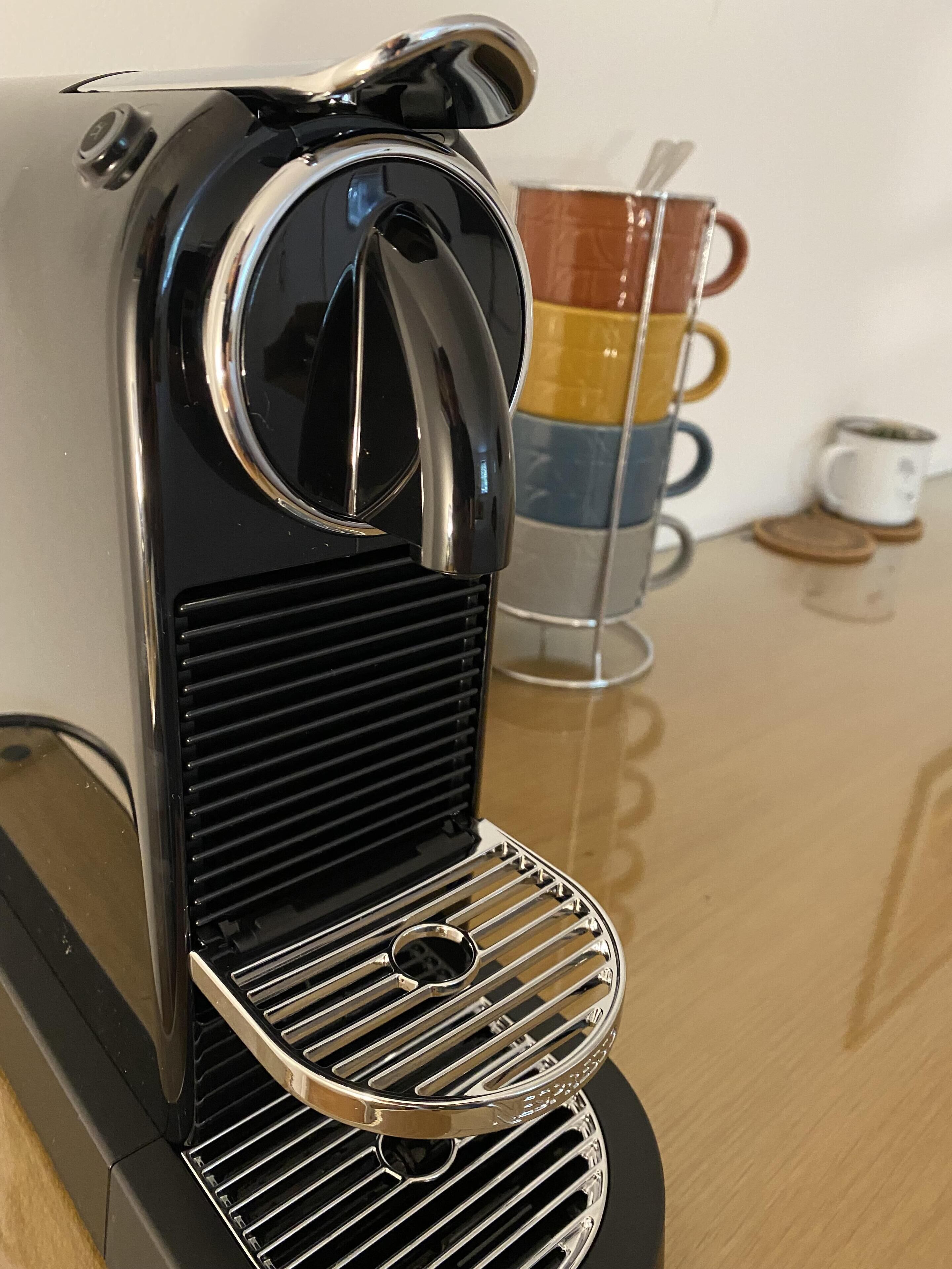 Una cafetera con un recipiente para goteo, un portavasos con varias tazas y un pequeño contenedor blanco sobre una superficie reflectante.