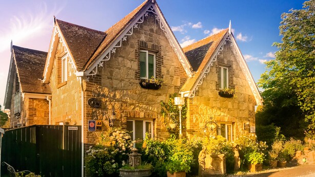 Un edificio de piedra tradicional con un tejado empinado, numerosas ventanas y un jardín bien cuidado con plantas en macetas y una fuente.