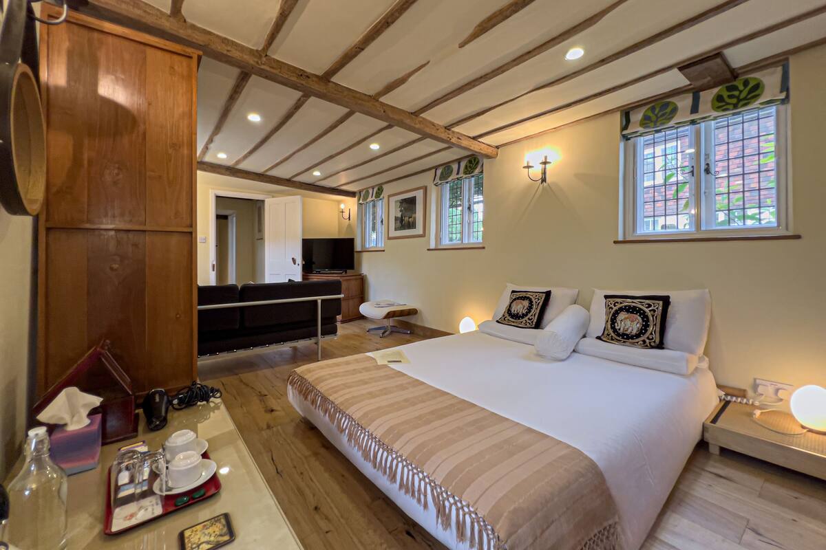 A hotel room with a large bed, wooden ceiling beams, a desk with a chair, a television, and a window with a view of greenery.