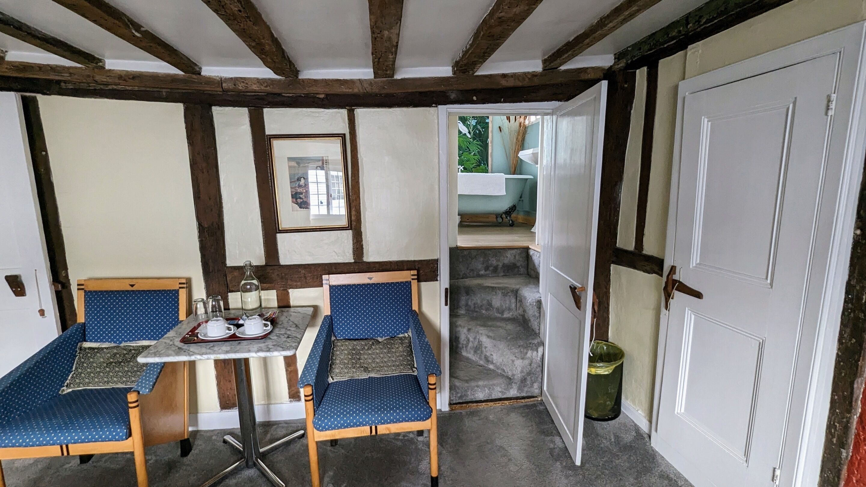 A room with a wooden ceiling, a small table with two chairs, a mirror, and a door leading to another room.