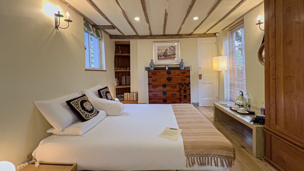 Een slaapkamer met een groot bed, houten balken in het plafond, een dressoir en een bureau met een lamp.