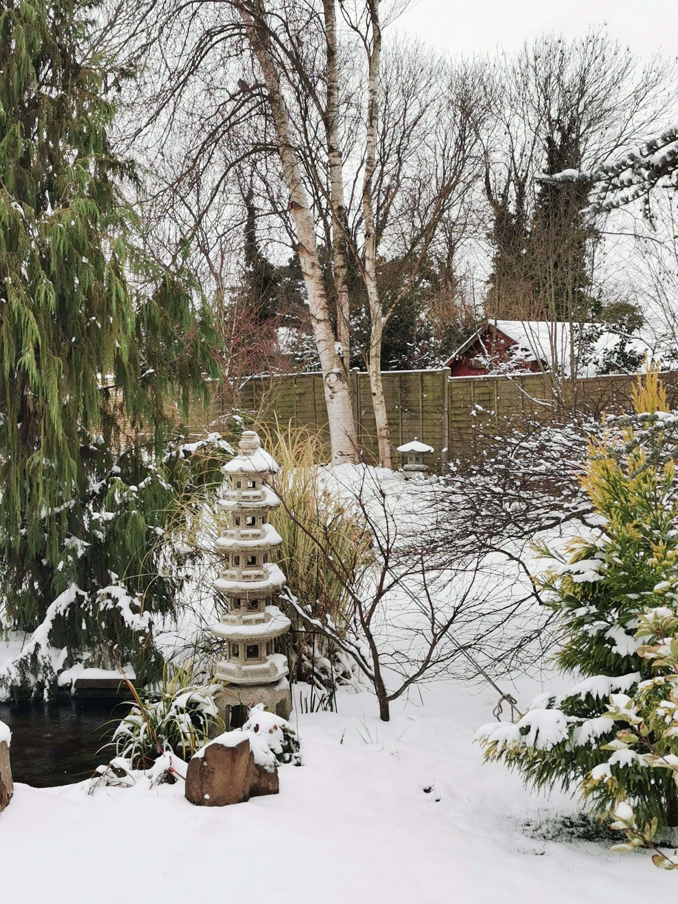 A garden with a stone pagoda, snow-covered trees, and a wooden fence.