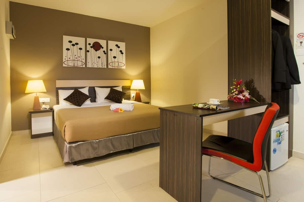 A hotel room with a bed, two bedside lamps, a desk with a chair, a small table with a cup and flowers, and a wardrobe.