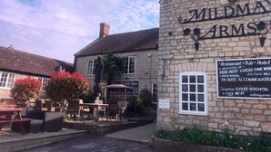 A stone building with a sign reading "MILD MAY ARM'S". It advertises as a restaurant, pub, and hotel offering a selection of real ales, ciders, and fine wines. It mentions award-winning a la carte menu and en-suite accommodation. The building has a car park for patrons only. - Mildmay Arms (Yeovil)
