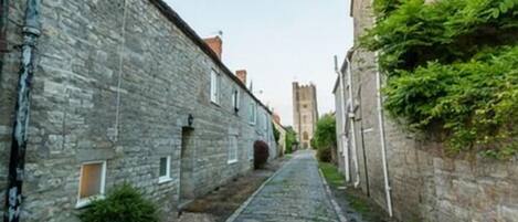 A narrow cobblestone street flanked by stone buildings with small windows and a tower in the distance.