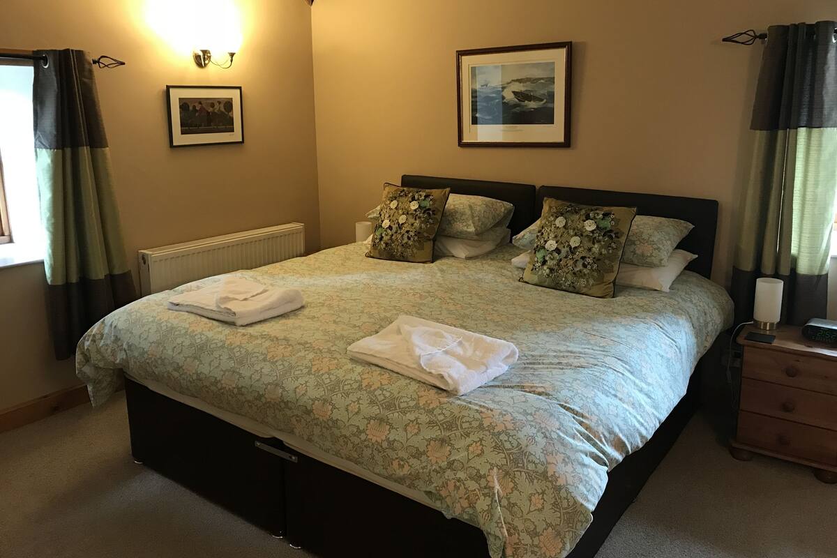 Cama bem arrumada com roupa de cama floral, mesa de cabeceira com abajur, quadro na parede e janela com cortinas.