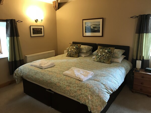 A neatly made bed with floral bedding, a bedside table with a lamp, a framed picture on the wall, and a window with curtains.