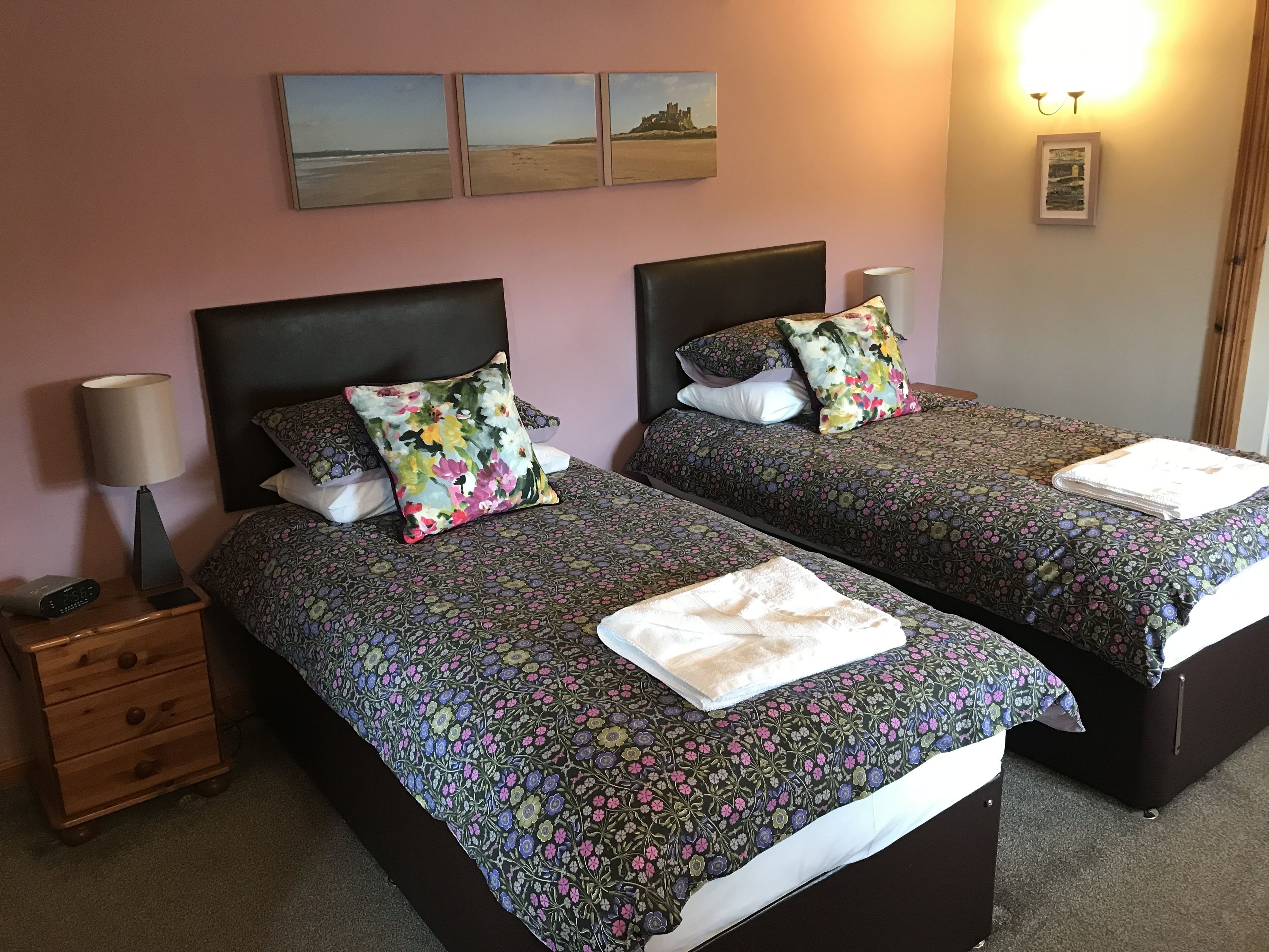 A hotel room with two beds, each with floral-patterned bedding and pillows, a bedside lamp, a nightstand, and a wall-mounted picture of a beach scene.