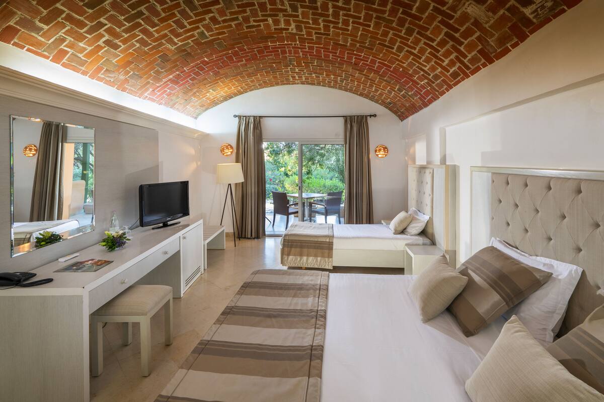A hotel room with a brick ceiling, a bed with multiple pillows, a desk with a television, and a view of the outdoors.
