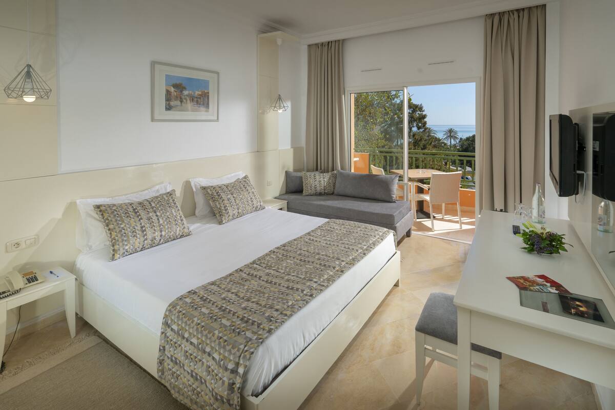 A hotel room with a large bed, a desk, and a balcony with a view of the sea.