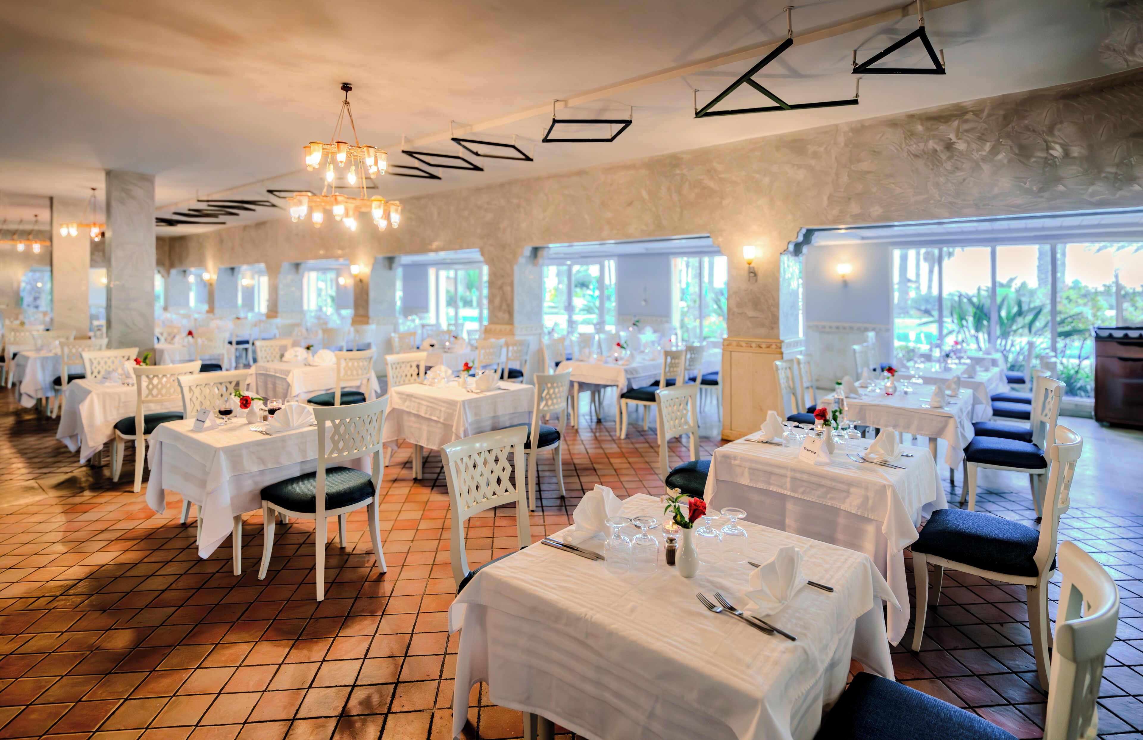 A restaurant with multiple tables and chairs, white tablecloths, and a tiled floor.