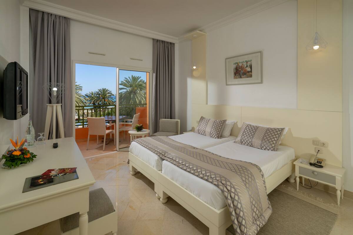 A hotel room with a large bed, a desk with a chair, a small table with a plant, and a balcony with a view of palm trees.