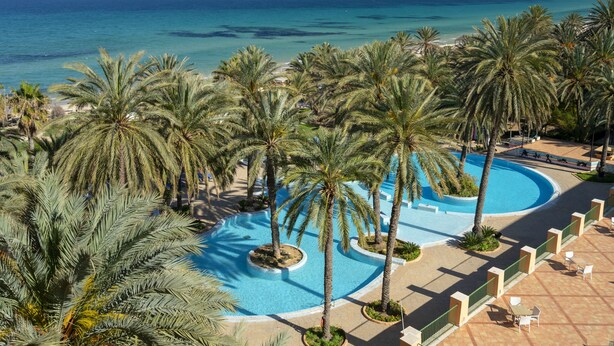Et resort med swimmingpool omgivet af palmer, med udsigt over en strand og havet.