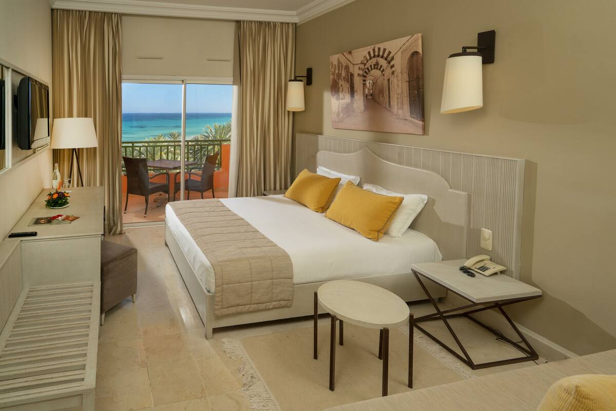A hotel room with a bed, a desk, a chair, a small table, a telephone, and a painting on the wall.
