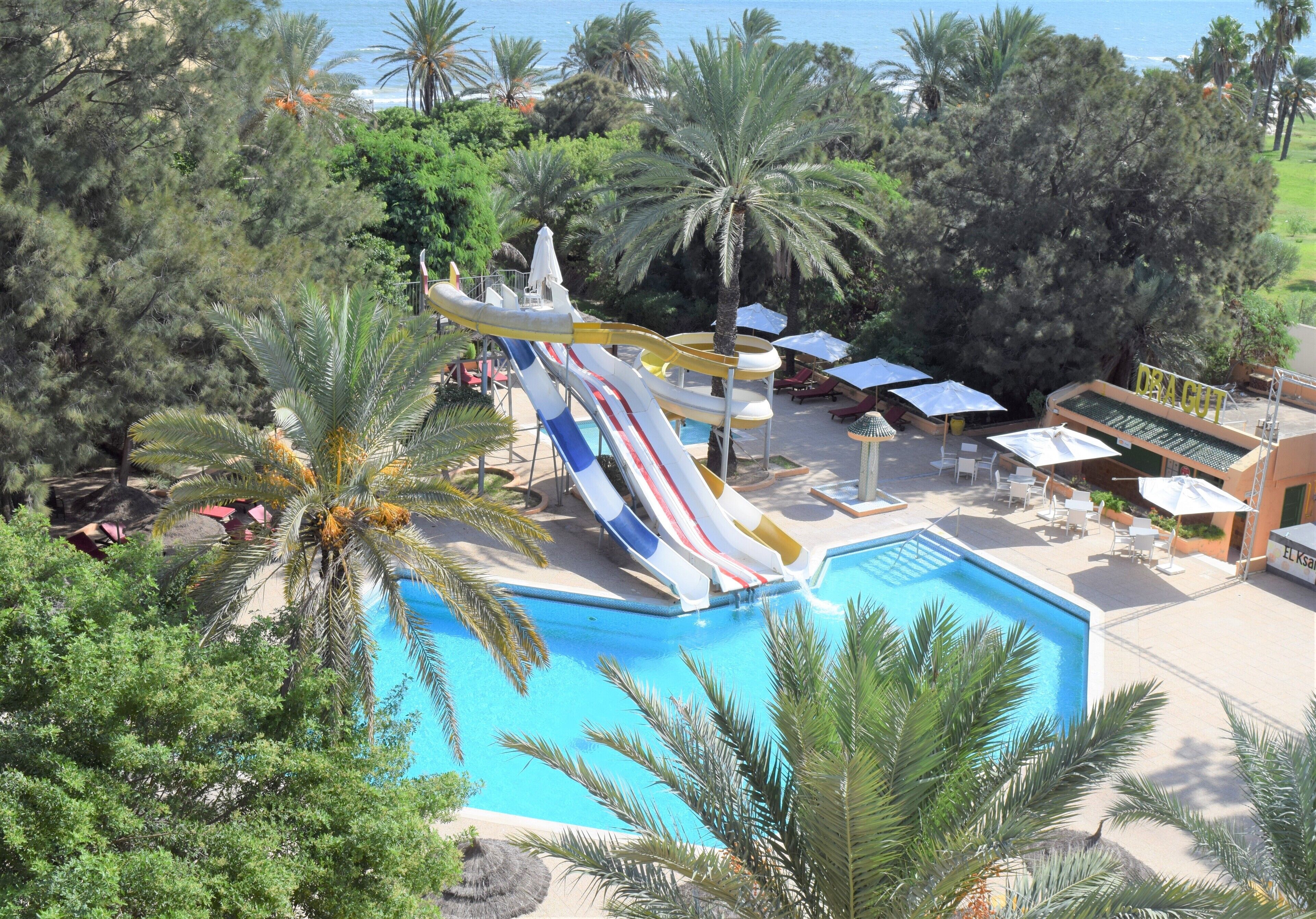 Ein Poolbereich mit Wasserrutsche, Palmen und Liegestühlen.