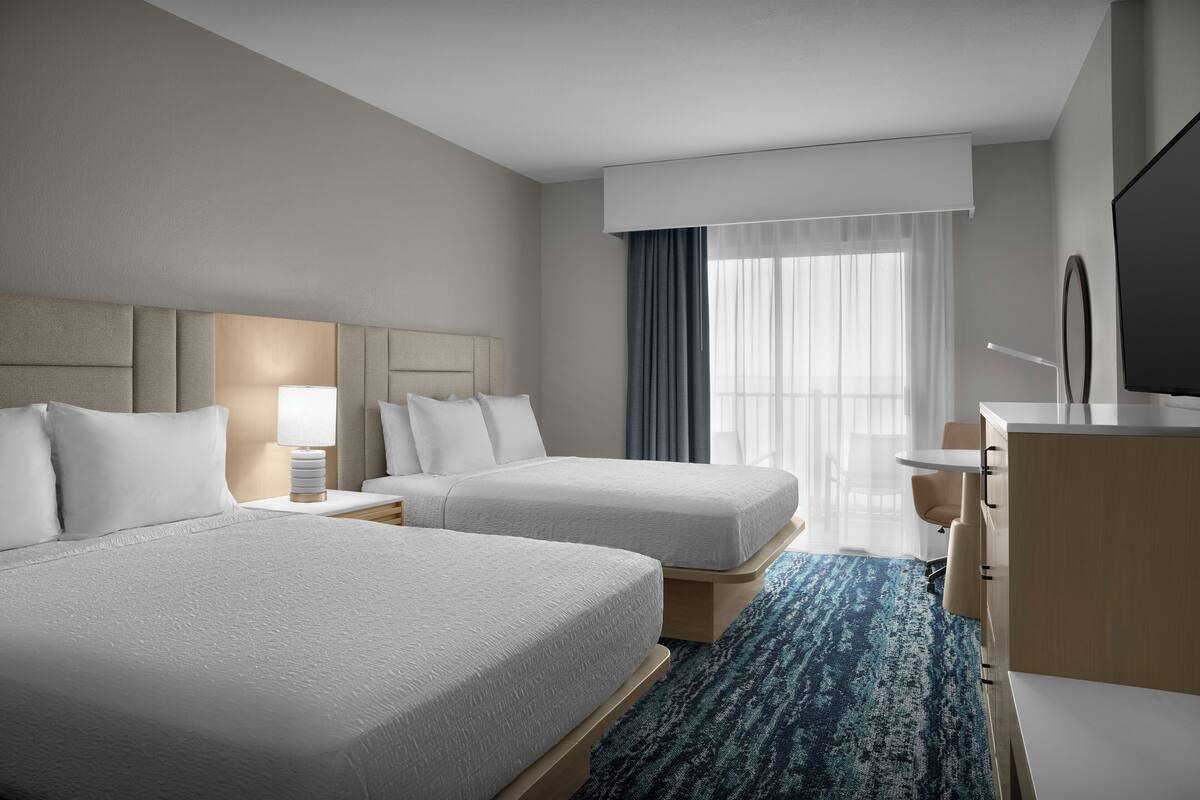 A hotel room with two beds, a desk, a TV, and a large window with curtains.
