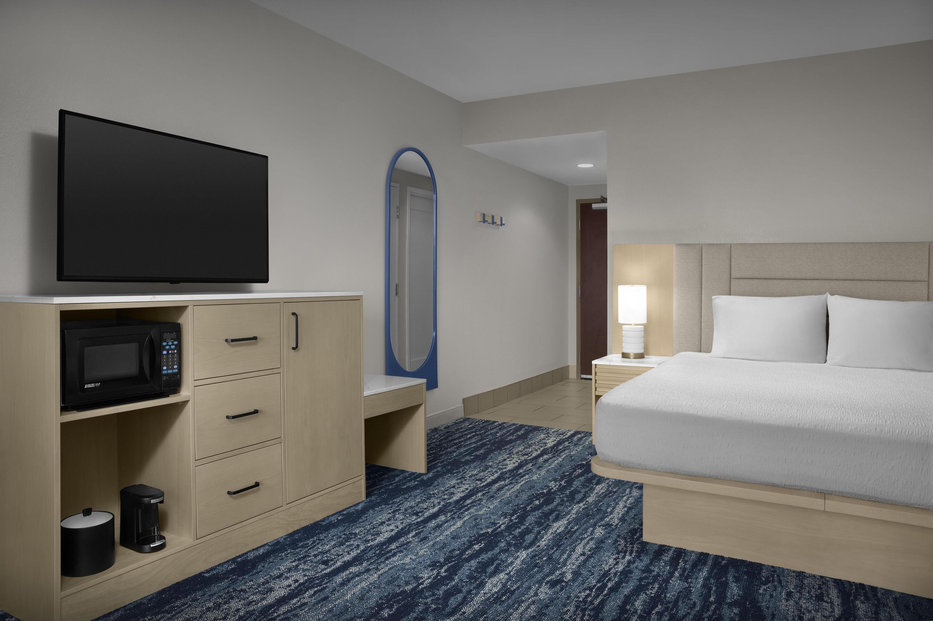A hotel room with a bed, a TV, a microwave, a coffee maker, a mirror, and a nightstand with a lamp.