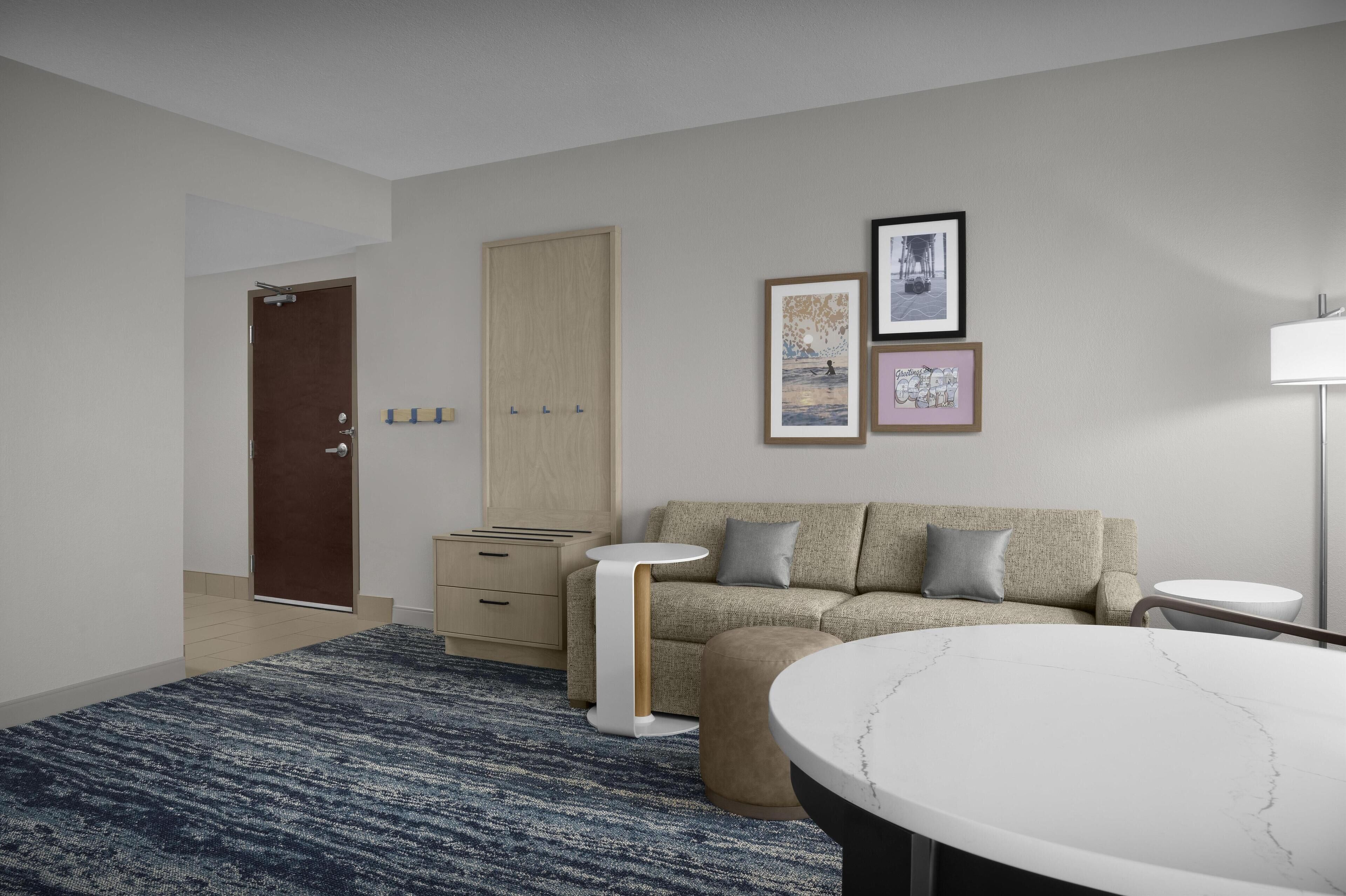 A modern hotel room with a sofa, a round table, a small side table, a wardrobe, and a door.