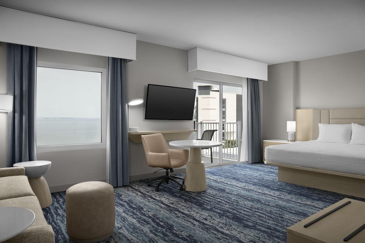 A hotel room with a bed, a desk, a chair, a TV, and a view of the outside.