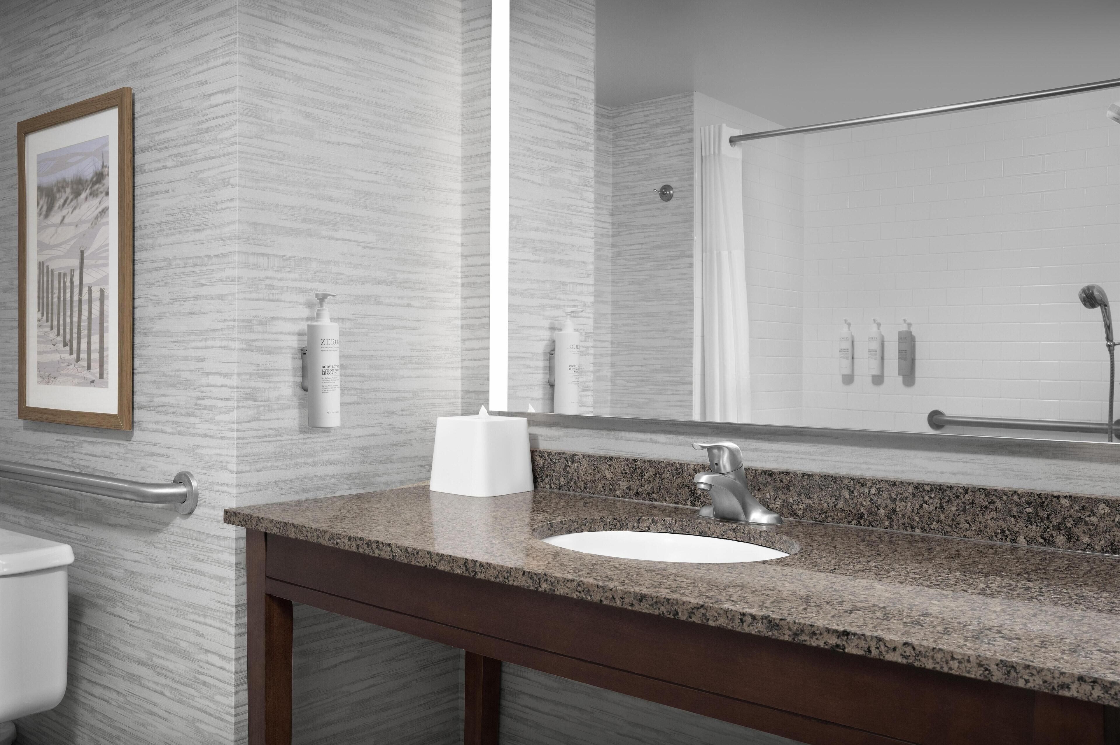 A bathroom with a granite countertop, a white sink, and a large mirror. There are toiletries on the counter and a framed picture on the wall.