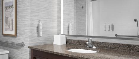 Un baño con encimera de granito, un lavabo blanco y un espejo grande. Hay artículos de aseo en la encimera y un cuadro en la pared.
